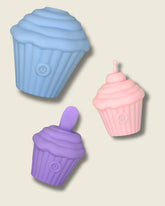 Three silicone cupcake-shaped discrete sex toys in blue, purple, and pink on a beige background.