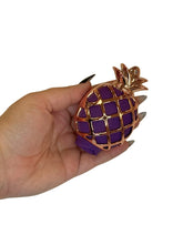 Hand holding a pineapple-shaped object with purple and gold design on a white background