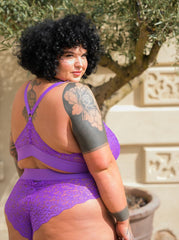 Curvy model wearing the Hallie lace bralette and high-waisted brief set in Passion Purple by Tutti Rouge, posing outdoors with back detail visible and floral lace design showcased.