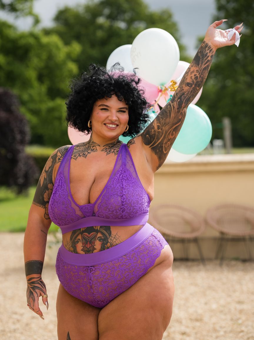 Smiling tattooed curve model wearing the Hallie Passion Purple lace bralette and matching briefs from Tutti Rouge, standing outdoors with balloons in the background.